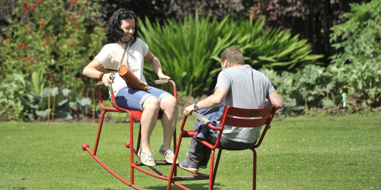 Een man en een vrouw zitten tegenover elkaar in een rode metalen schommelstoel in de tuin van Arboretum Kalmthout. Ze hebben er duidelijk plezier in. De zon schijnt.