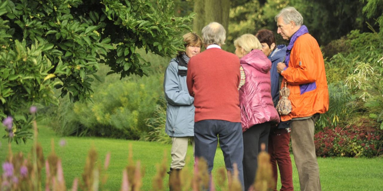 Foto van een park, op de achtergrond een klein groepje bezoekers op het gras. Een gids vertelt hen iets, ze luisteren aandachtig naar haar.