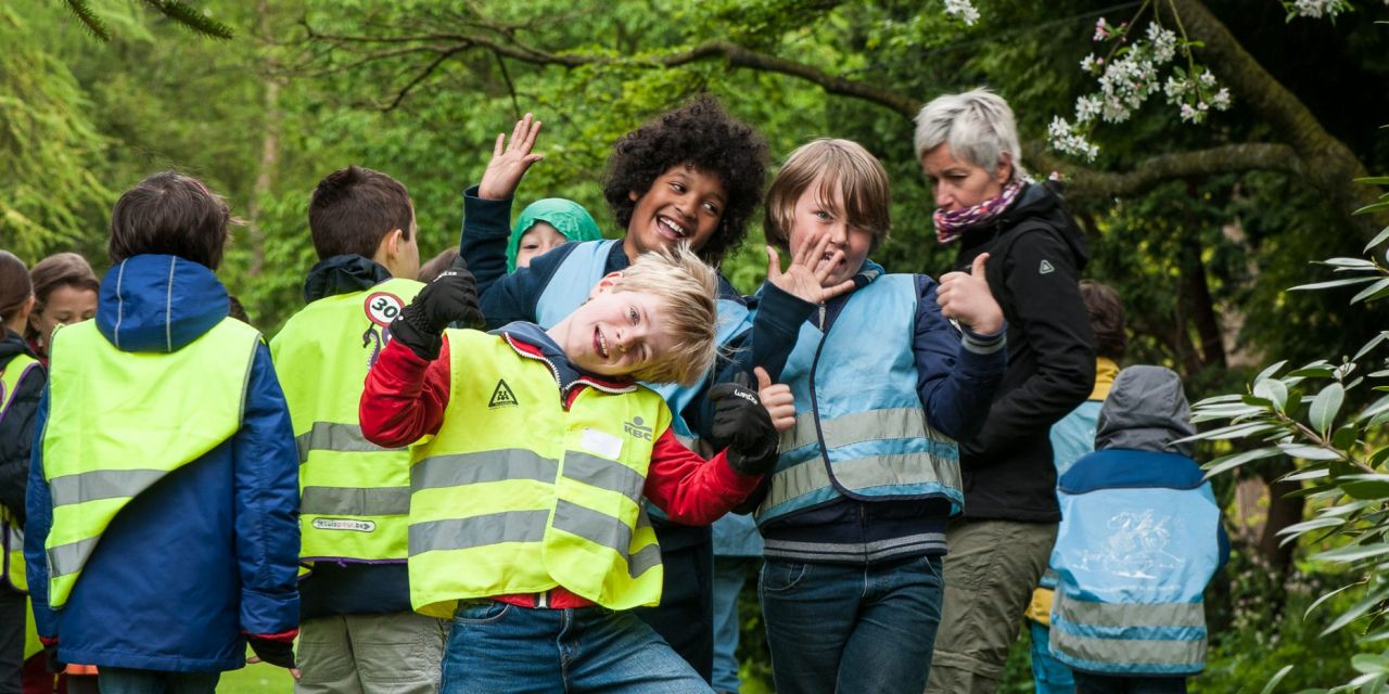 Een groep schoolkinderen met hun juf tussen de bomen van Arboretum Kalmthout. Drie jongens poseren voor de foto, zwaaien of steken vrolijk hun duim op.