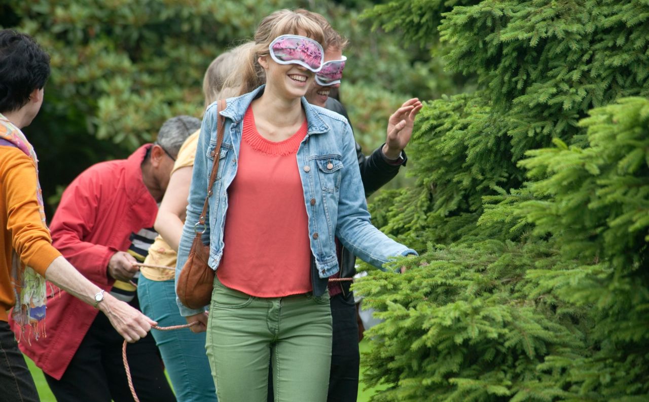 Terra Nova blinddoekwandeling in Arboretum Kalmthout, enkele bezoekers maken een wandeling met een slaapmaskertje en voelen aan een struik. Een gids leidt hen rond met een touw.